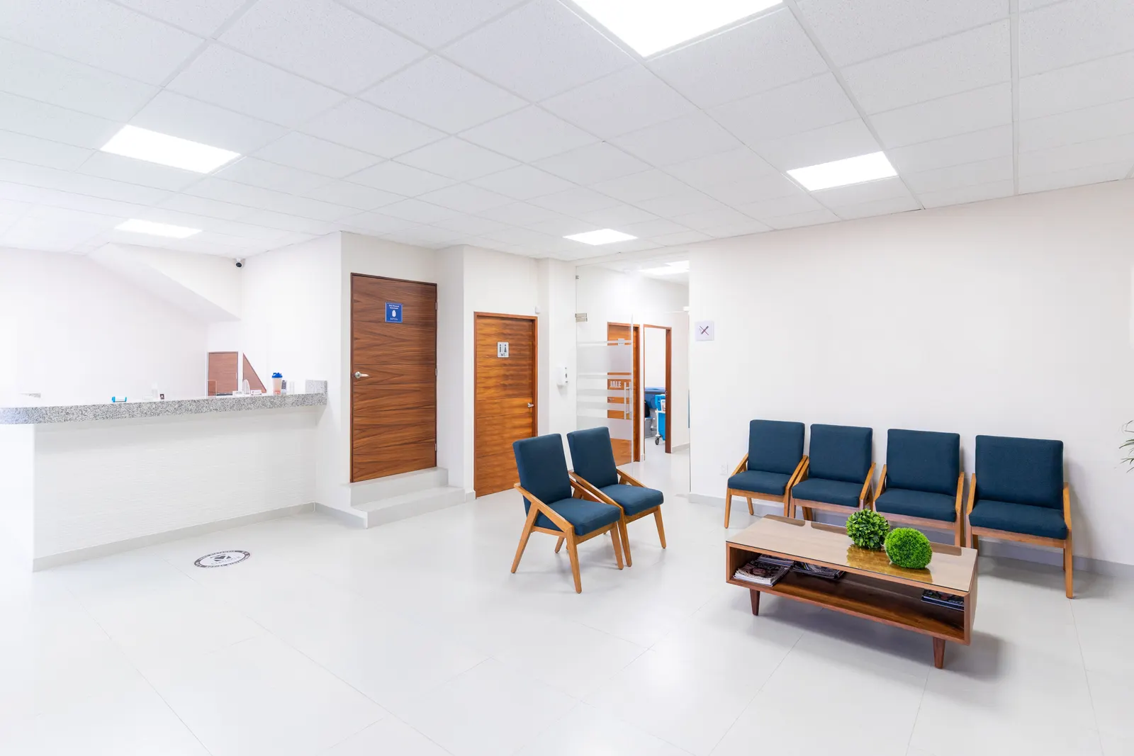 Sala de espera de clínica com cadeiras azuis, balcão de recepção e portas de madeira, sem pessoas